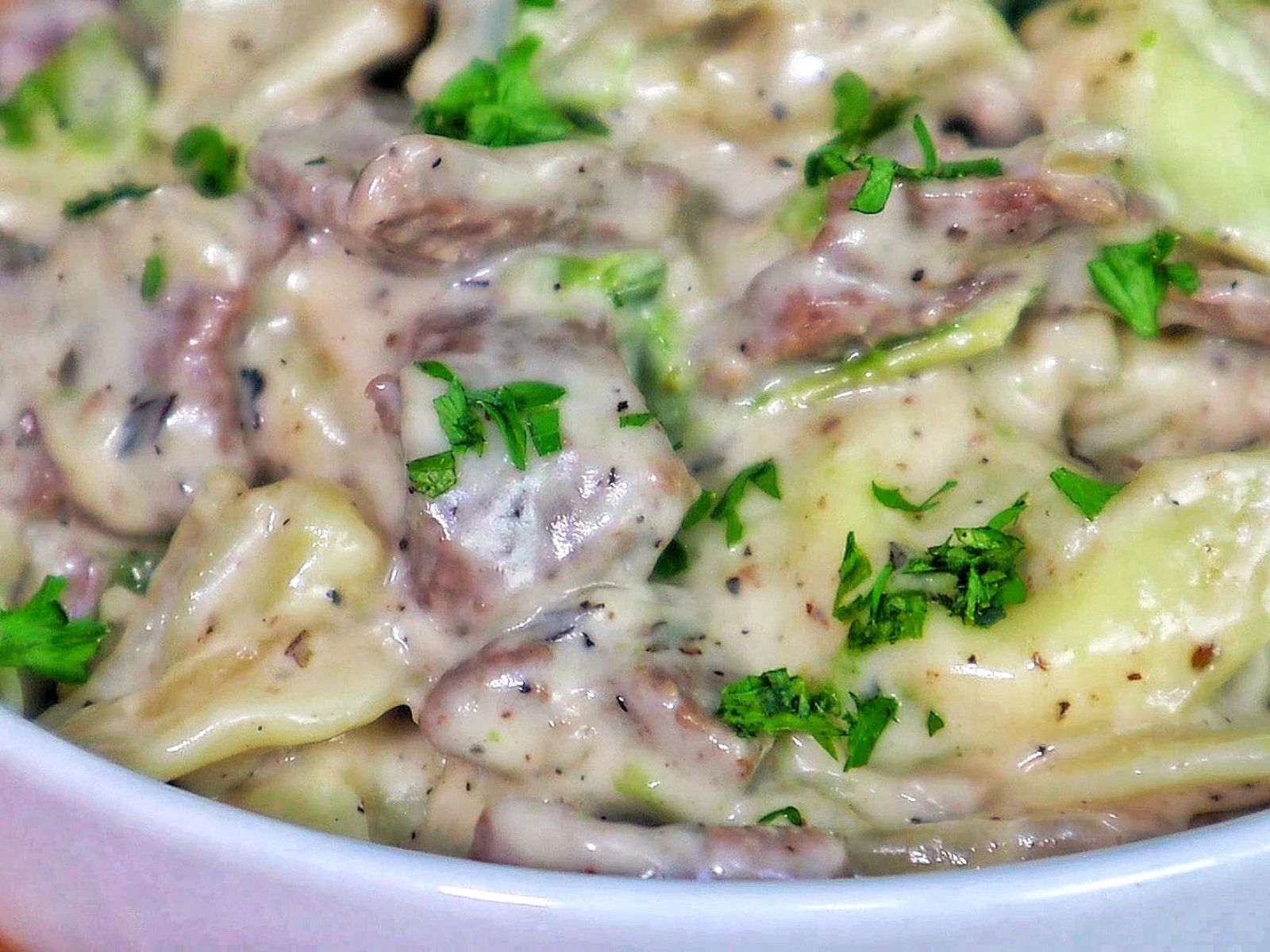 A creamy bowl of cheesesteak tortellini, with thinly sliced beef, cheese-filled tortellini pasta, and green bell peppers coated in a rich, seasoned cream sauce. Garnished with chopped parsley for a fresh finish.