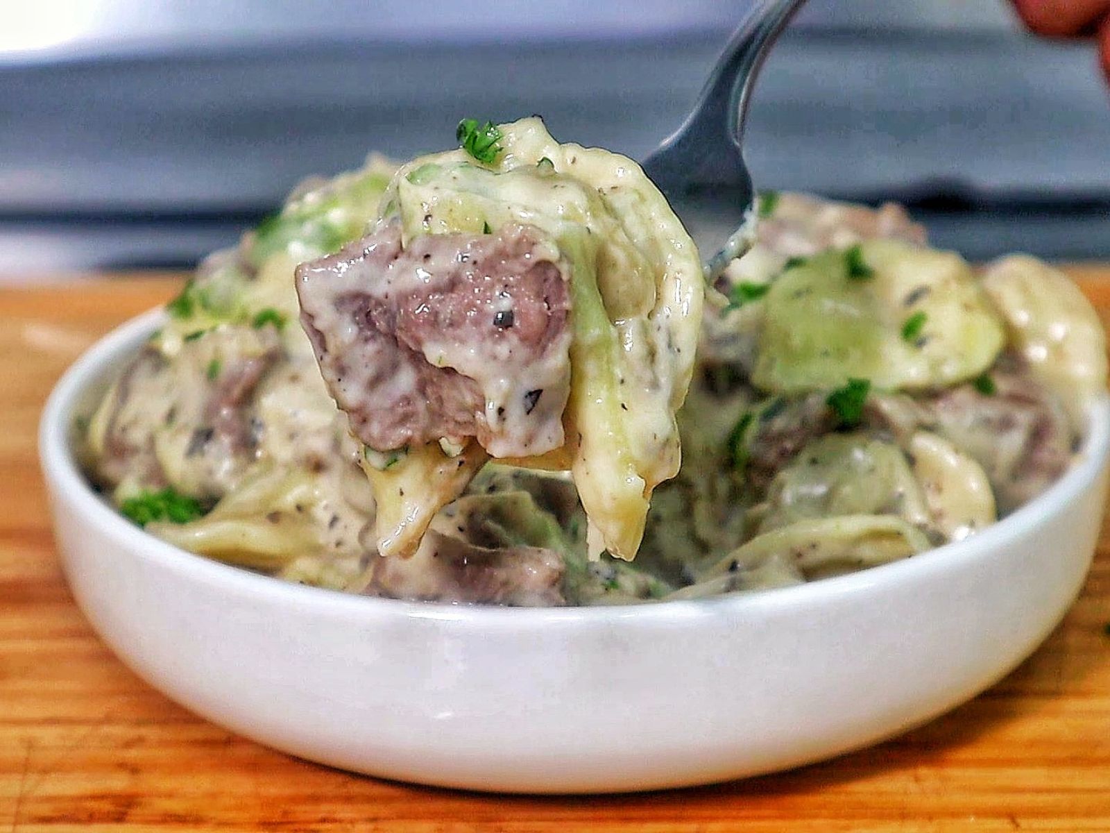 A creamy bowl of cheesesteak tortellini, with thinly sliced beef, cheese-filled tortellini pasta, and green bell peppers coated in a rich, seasoned cream sauce. Garnished with chopped parsley for a fresh finish.