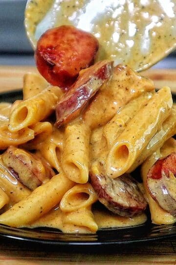 A creamy serving of cheesy ranch chicken and sausage pasta on a black plate. The dish features tender penne pasta coated in a ranch-seasoned cheese sauce, with seared chicken chunks and slices of smoked sausage.