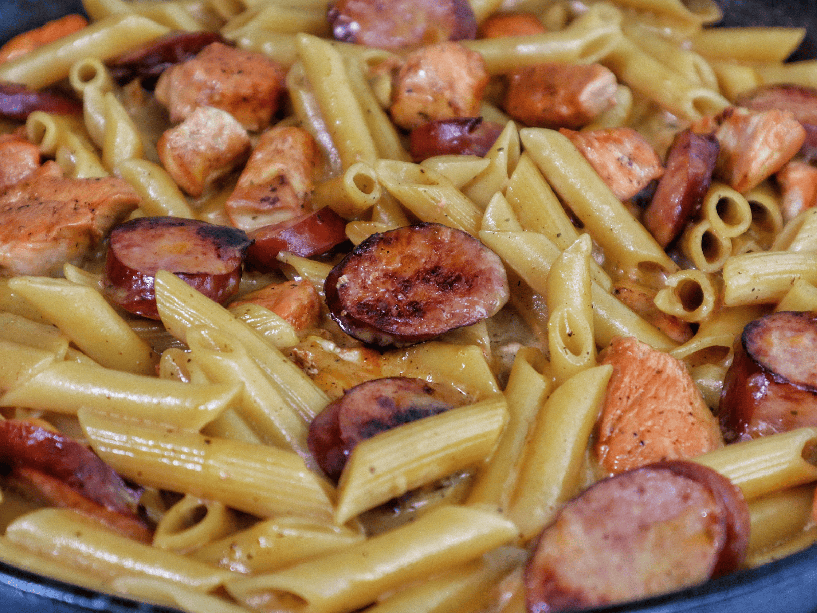 A creamy serving of cheesy ranch chicken and sausage pasta on a black plate. The dish features tender penne pasta coated in a ranch-seasoned cheese sauce, with seared chicken chunks and slices of smoked sausage.