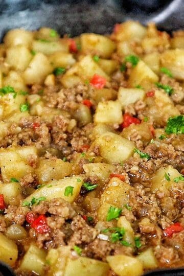 A cast iron skillet filled with a mixture of ground beef, tender potato chunks, red bell peppers, and chopped parsley, all simmered in a savory, seasoned sauce.