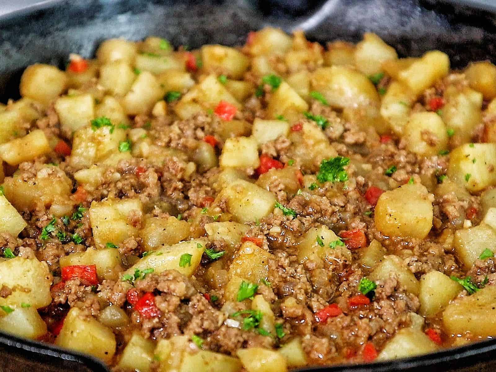 A cast iron skillet filled with a mixture of ground beef, tender potato chunks, red bell peppers, and chopped parsley, all simmered in a savory, seasoned sauce.
