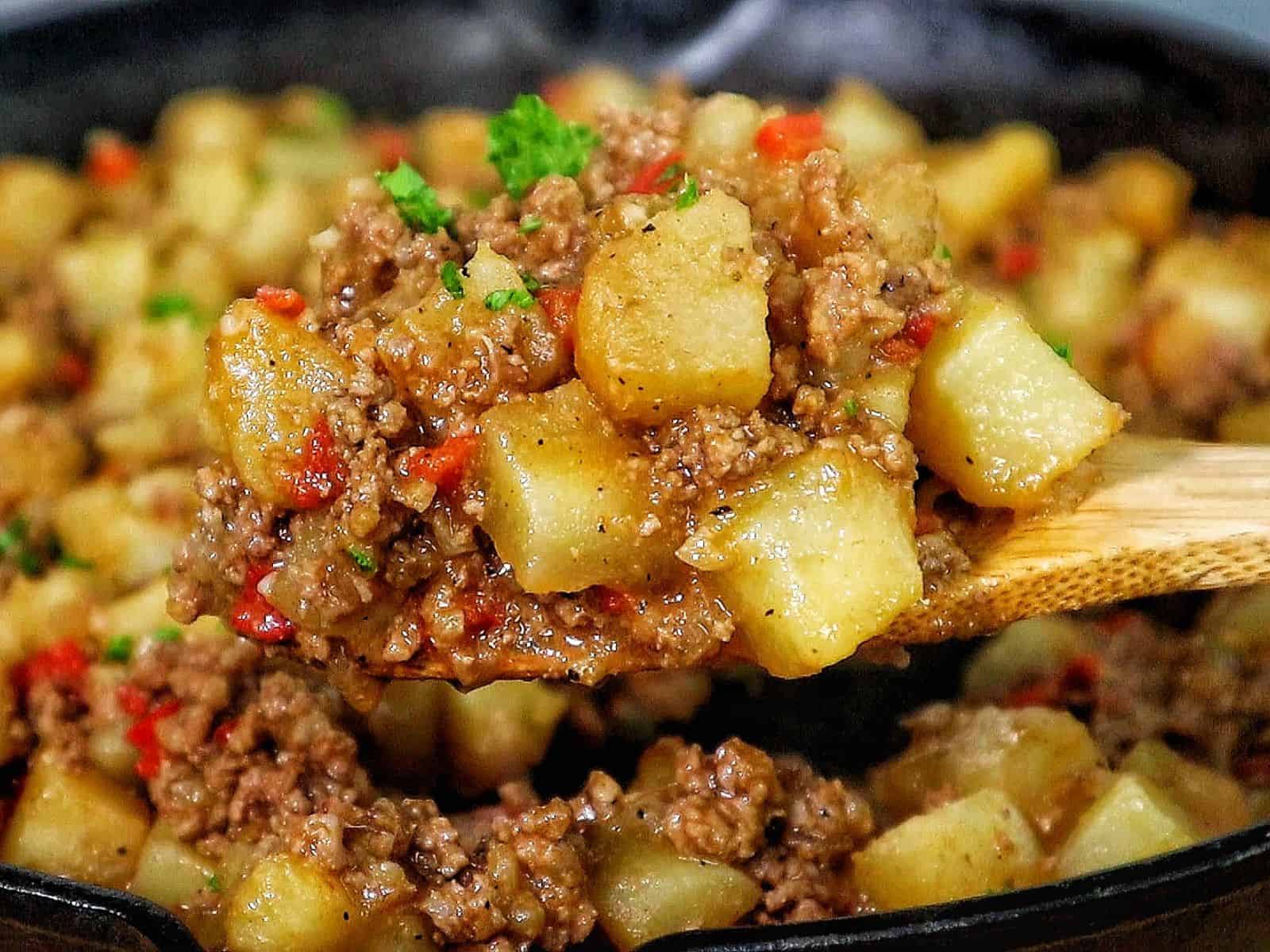 A cast iron skillet filled with a mixture of ground beef, tender potato chunks, red bell peppers, and chopped parsley, all simmered in a savory, seasoned sauce.