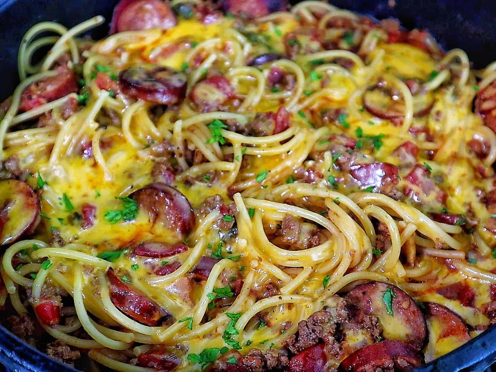Cheesy cowboy spaghetti made with spaghetti noodles, slices of polish sausage, ground beef, diced tomatoes, and melted cheddar cheese, garnished with fresh parsley.