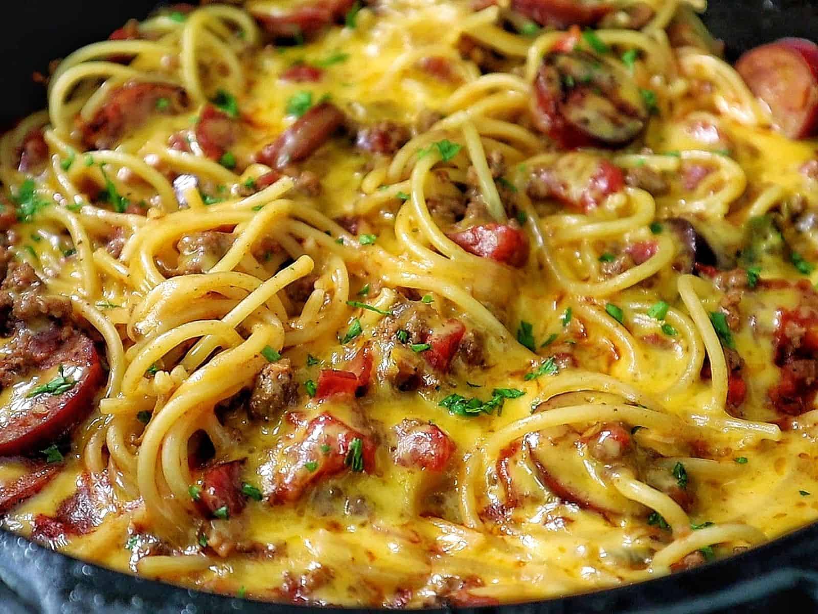 Cheesy cowboy spaghetti made with spaghetti noodles, slices of polish sausage, ground beef, diced tomatoes, and melted cheddar cheese, garnished with fresh parsley.