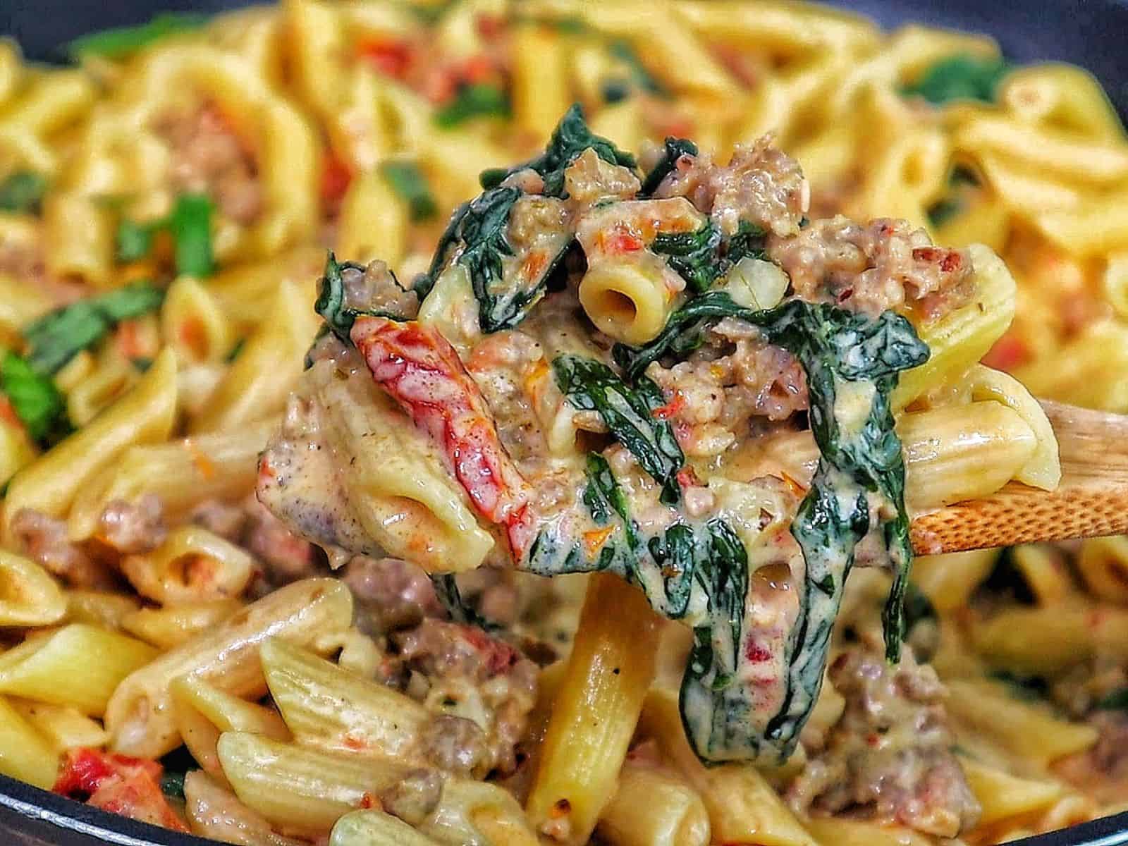 A skillet filled with creamy Tuscan sausage pasta made with penne, Italian sausage, sun-dried tomatoes, and fresh spinach, coated in a rich cream sauce and garnished with fresh basil.