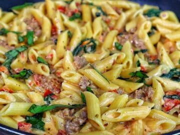 A skillet filled with creamy Tuscan sausage pasta made with penne, Italian sausage, sun-dried tomatoes, and fresh spinach, coated in a rich cream sauce and garnished with fresh basil.