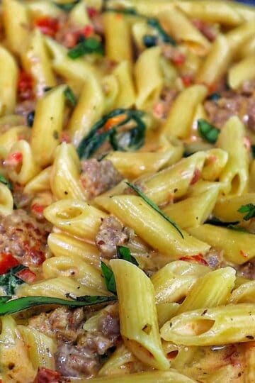 A skillet filled with creamy Tuscan sausage pasta made with penne, Italian sausage, sun-dried tomatoes, and fresh spinach, coated in a rich cream sauce and garnished with fresh basil.