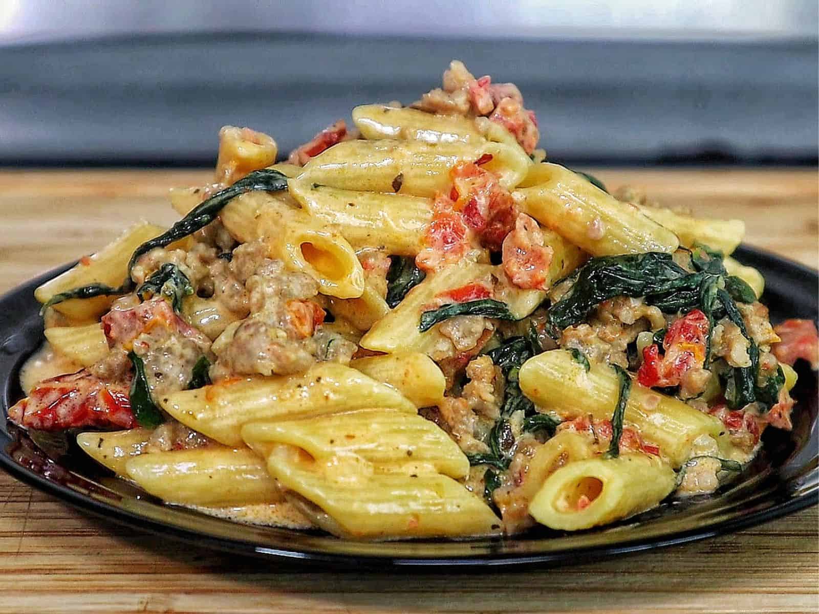 A skillet filled with creamy Tuscan sausage pasta made with penne, Italian sausage, sun-dried tomatoes, and fresh spinach, coated in a rich cream sauce and garnished with fresh basil.