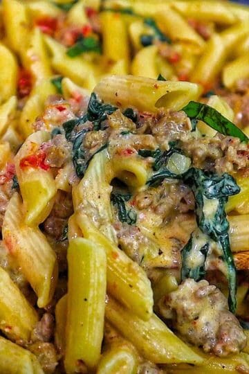 A skillet filled with creamy Tuscan sausage pasta made with penne, Italian sausage, sun-dried tomatoes, and fresh spinach, coated in a rich cream sauce and garnished with fresh basil.
