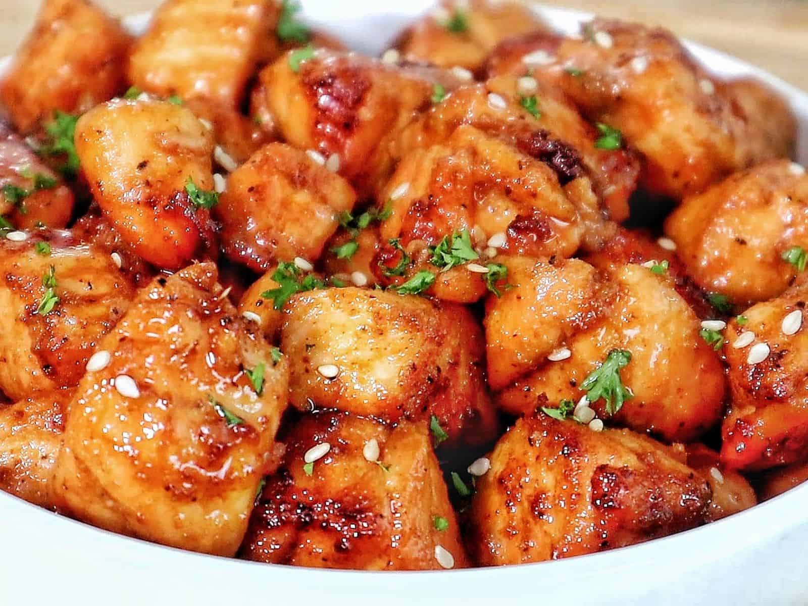 A white bowl filled with golden brown hot honey salmon bites, garnished with sesame seeds and fresh parsley.