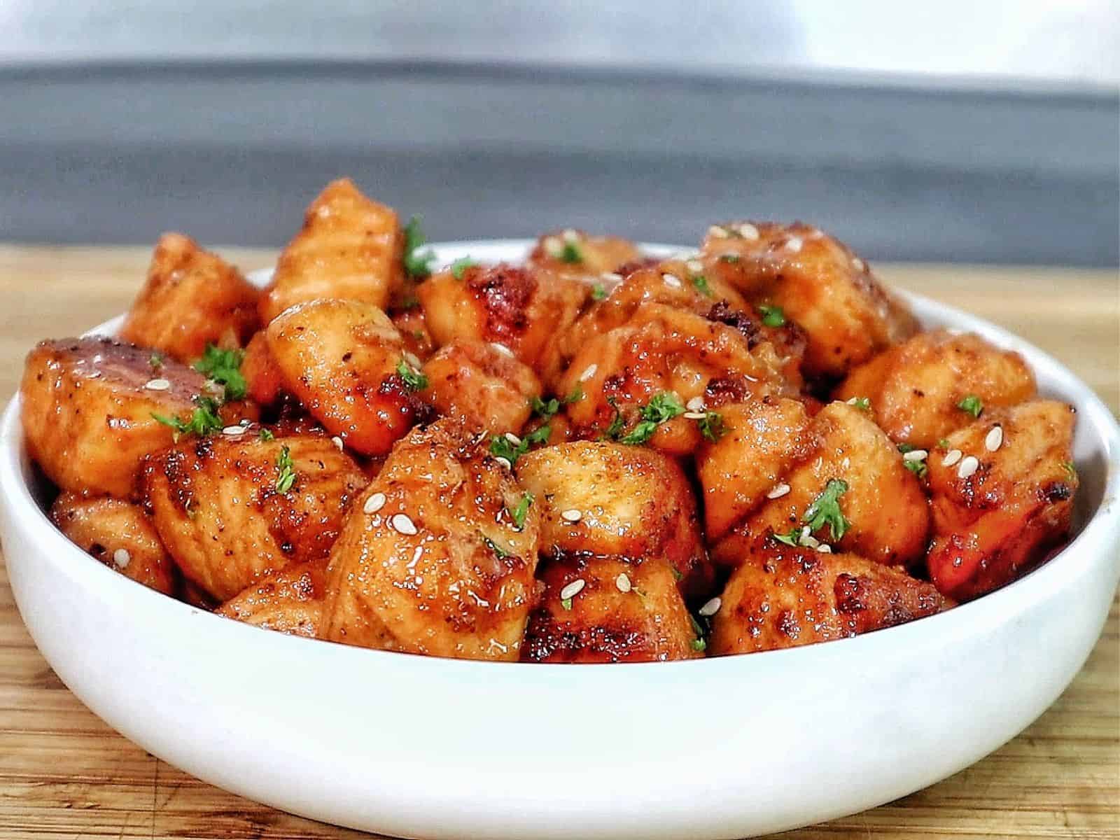 A white bowl filled with golden brown hot honey salmon bites, garnished with sesame seeds and fresh parsley.