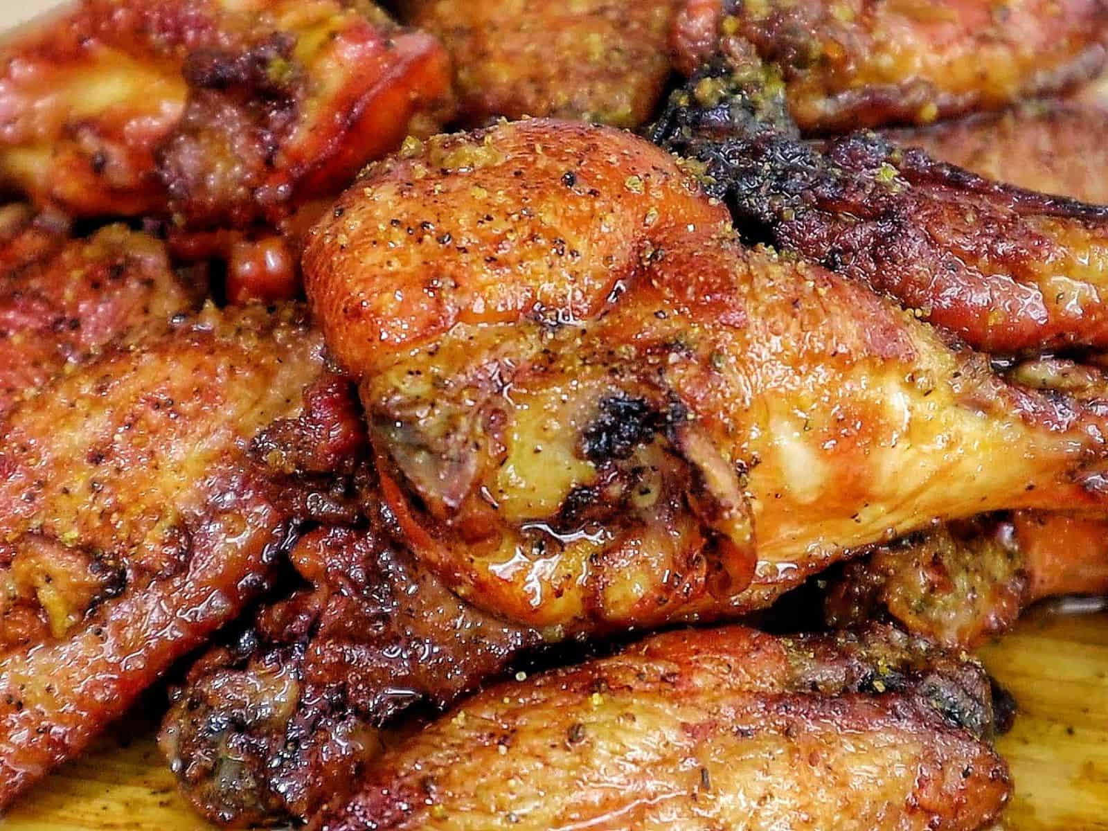 A close-up of golden-brown smoked chicken wings piled on a wooden cutting board, seasoned with a wet lemon pepper sauce from the grill.