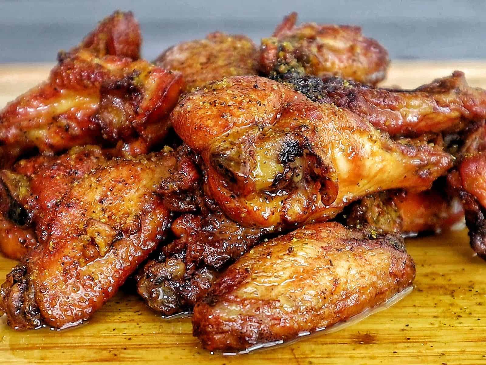A close-up of golden-brown smoked chicken wings piled on a wooden cutting board, seasoned with a wet lemon pepper sauce from the grill.