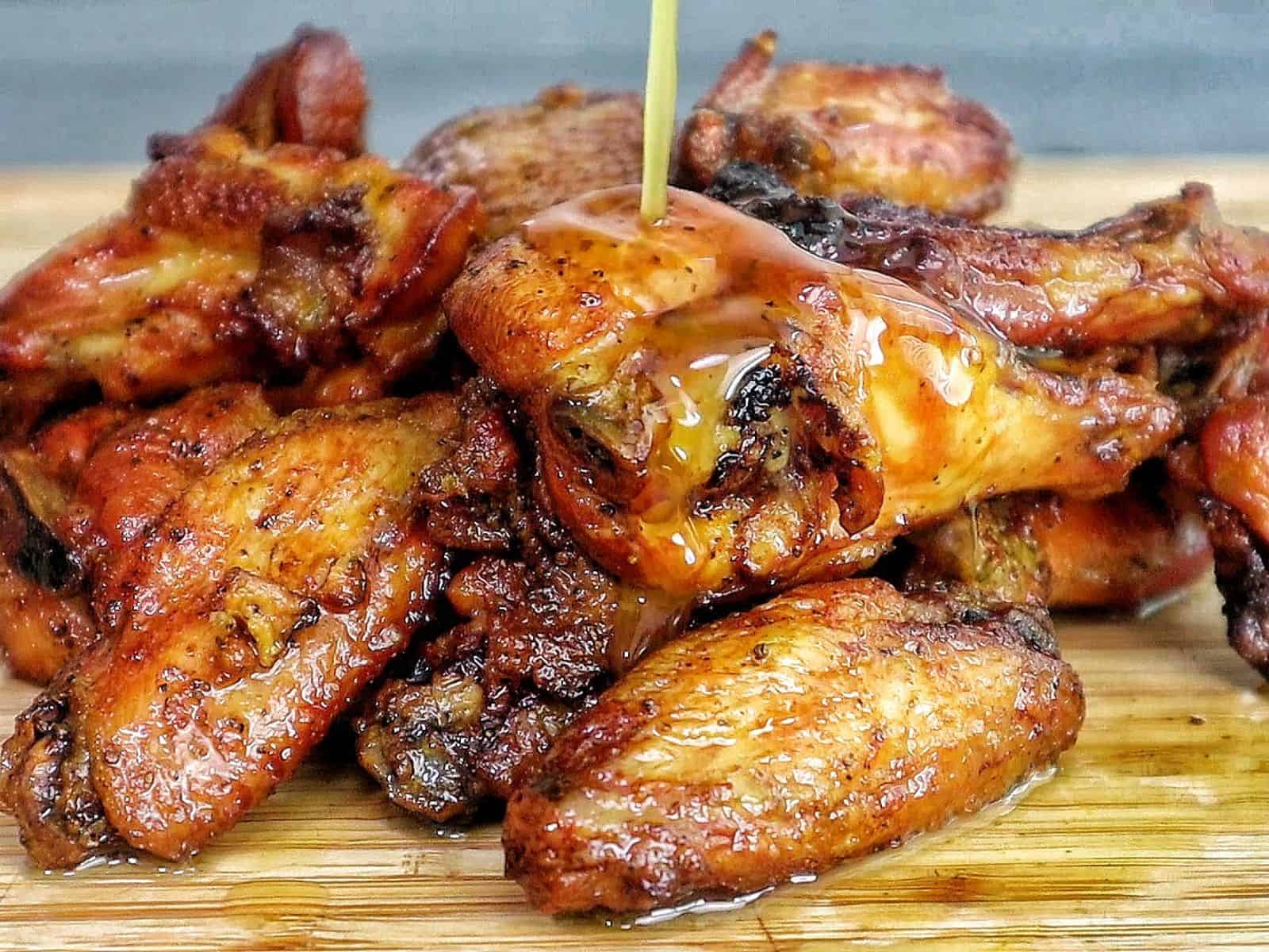 A close-up of golden-brown smoked chicken wings piled on a wooden cutting board, seasoned with a wet lemon pepper sauce from the grill.