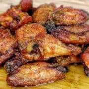 A close-up of golden-brown smoked chicken wings piled on a wooden cutting board, seasoned with a wet lemon pepper sauce from the grill.