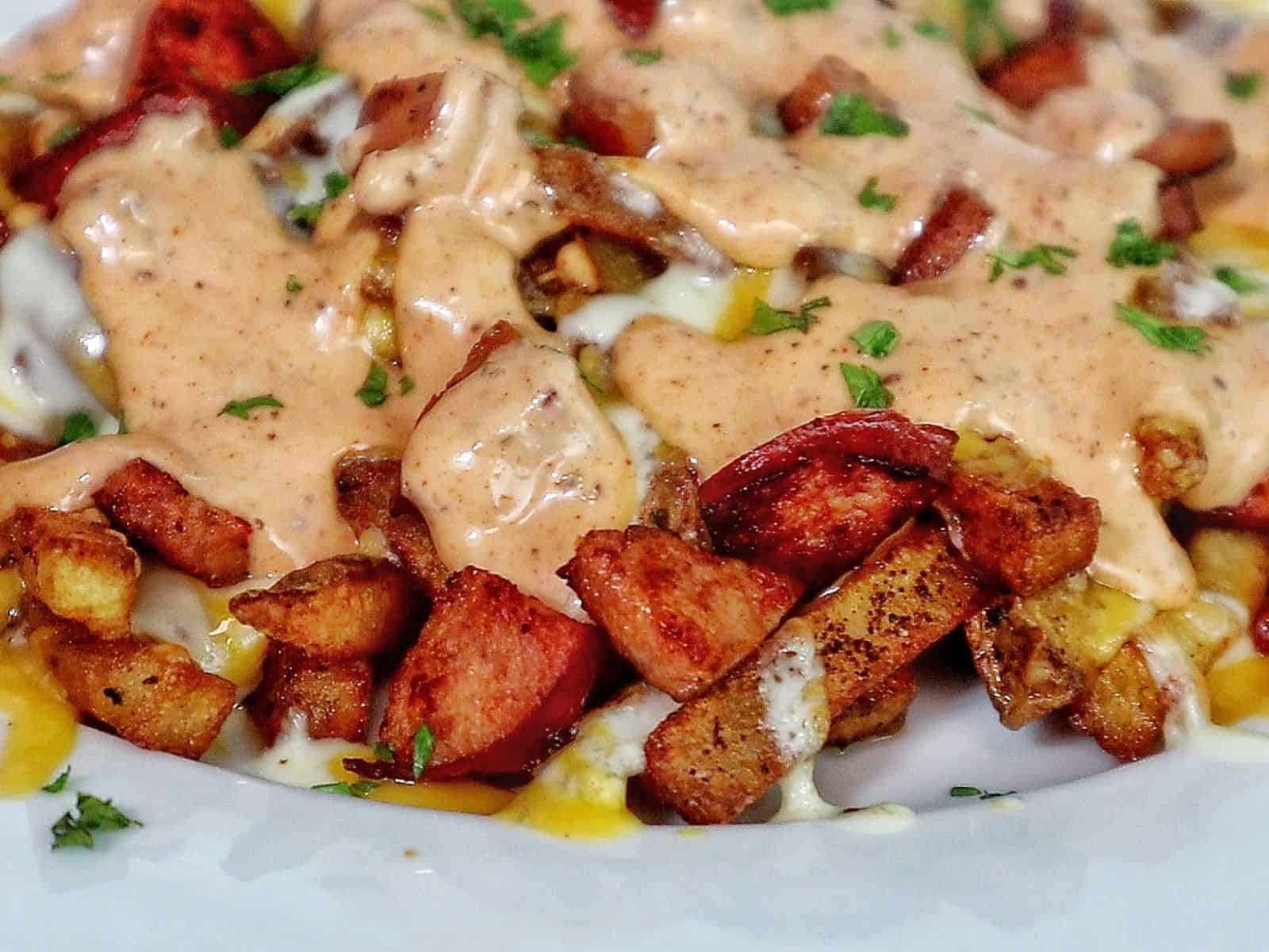 Golden, crispy French fries topped with melted cheese, Cajun sausage, and drizzled with spicy ranch-style sauce, garnished with fresh herbs.
