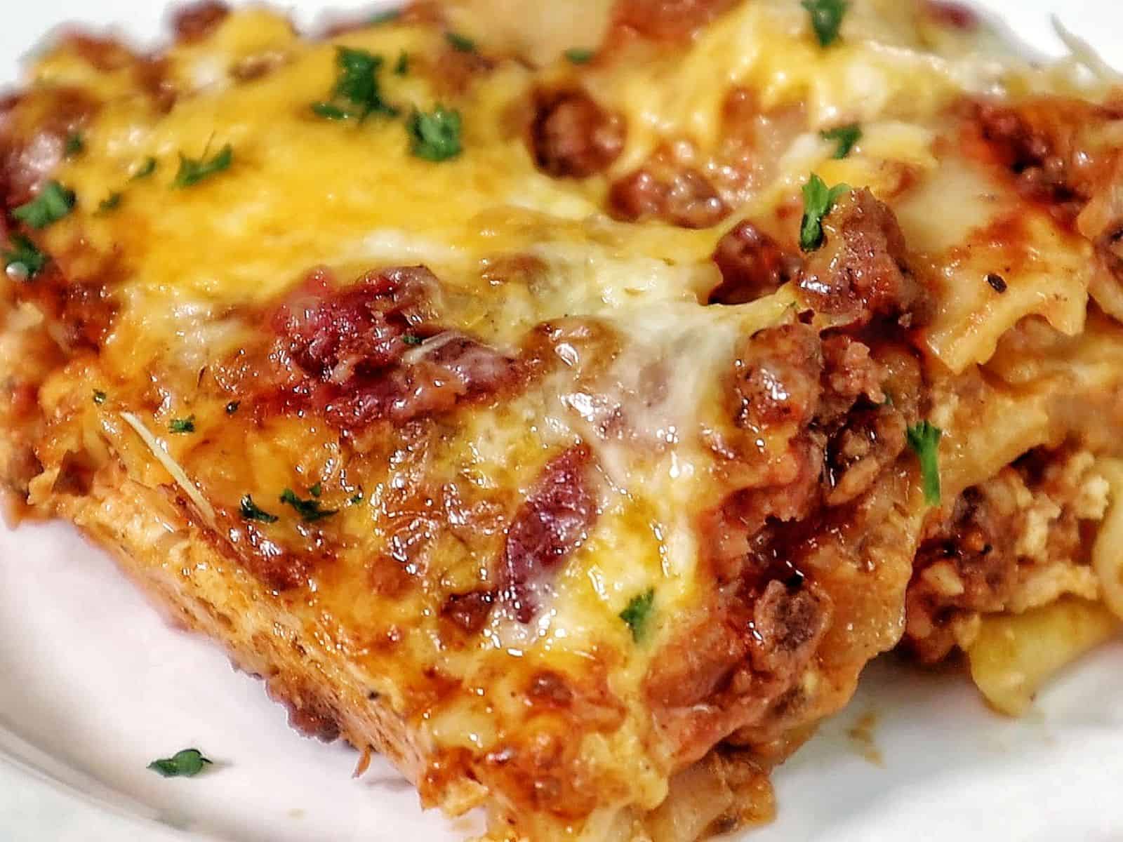 A slice of parmesan garlic bacon cheeseburger lasagna layered with pasta, seasoned beef, crispy bacon, rich tomato sauce, and melted cheese, served on a white plate.