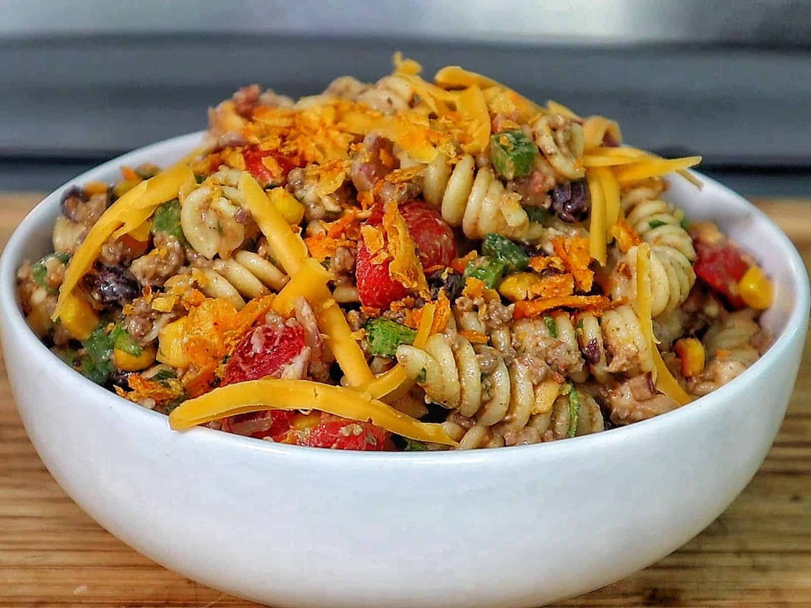A bowl filled with taco pasta made with rotini noodles, ground beef, black beans, corn, tomatoes, green peppers, and topped with shredded cheddar cheese and crushed chips.