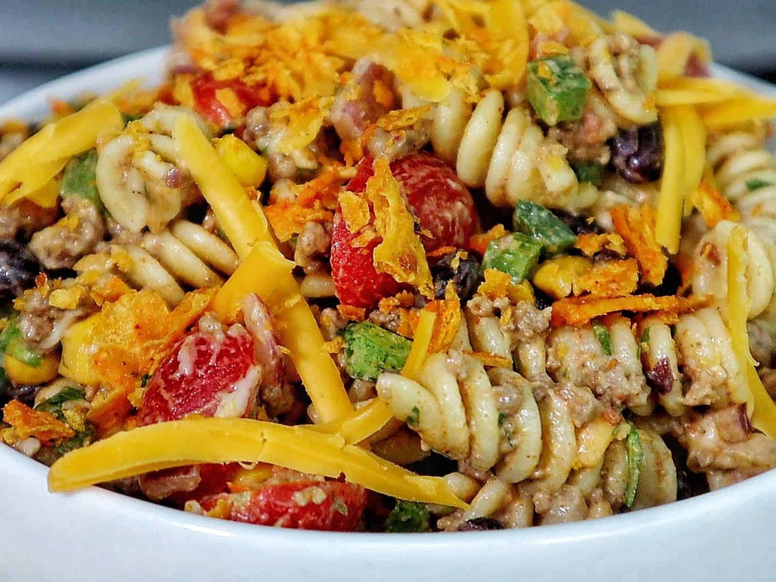 A bowl filled with taco pasta made with rotini noodles, ground beef, black beans, corn, tomatoes, green peppers, and topped with shredded cheddar cheese and crushed chips.
