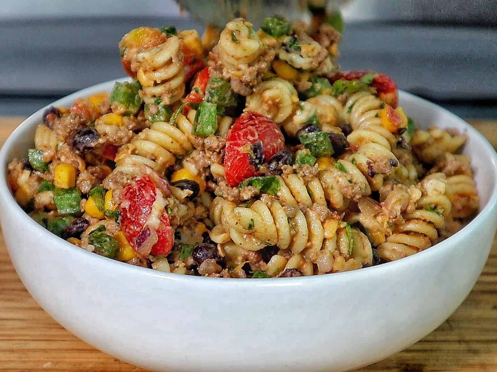 A bowl filled with taco pasta made with rotini noodles, ground beef, black beans, corn, tomatoes, green peppers, and topped with shredded cheddar cheese and crushed chips.