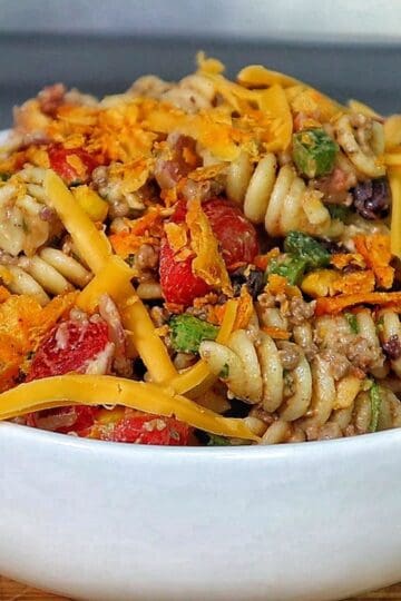 A bowl filled with taco pasta made with rotini noodles, ground beef, black beans, corn, tomatoes, green peppers, and topped with shredded cheddar cheese and crushed chips.