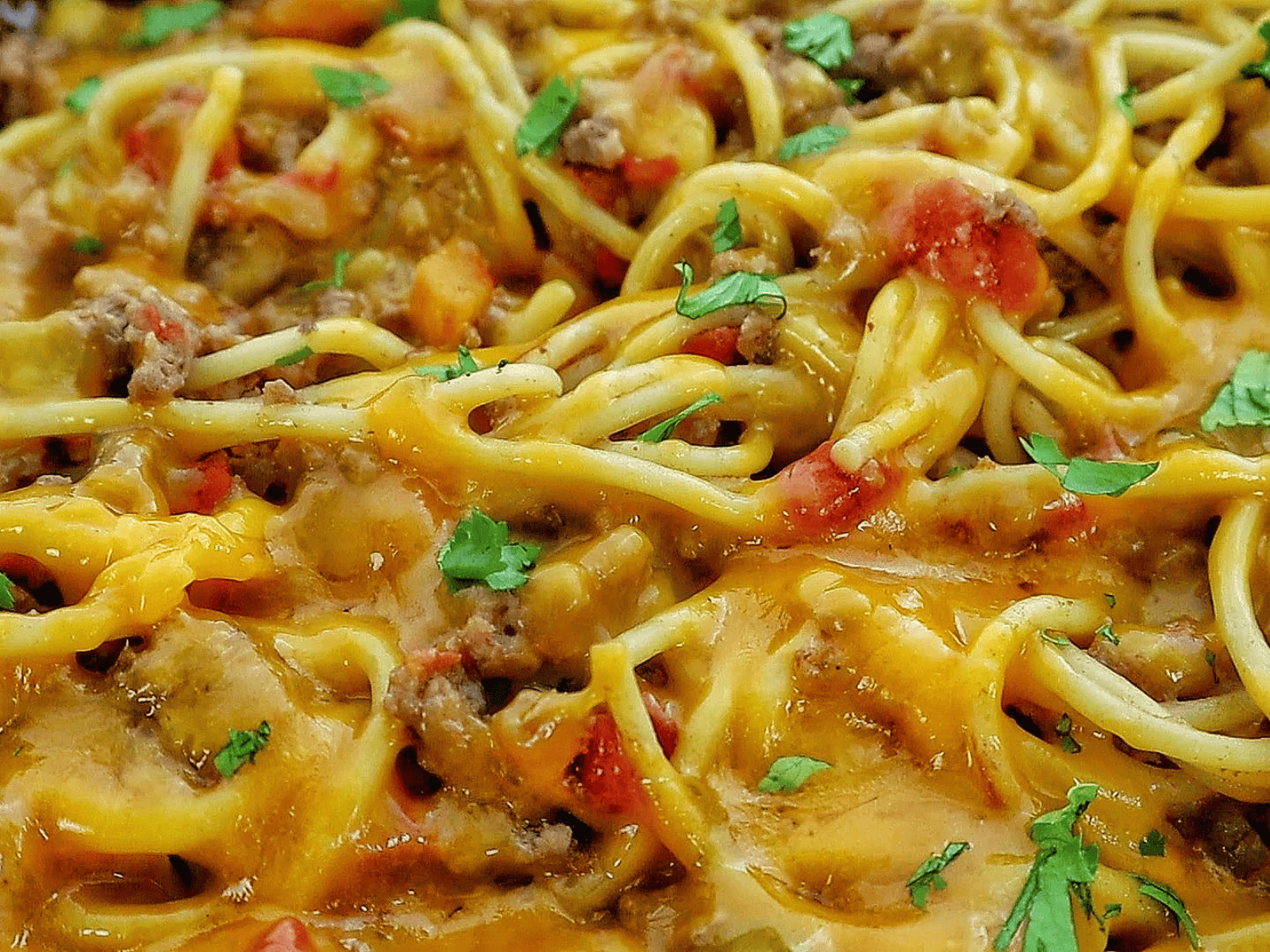 A skillet filled with cheesy taco spaghetti, featuring seasoned ground beef, spaghetti noodles, melted cheddar cheese, diced tomatoes, and garnished with chopped fresh parsley.