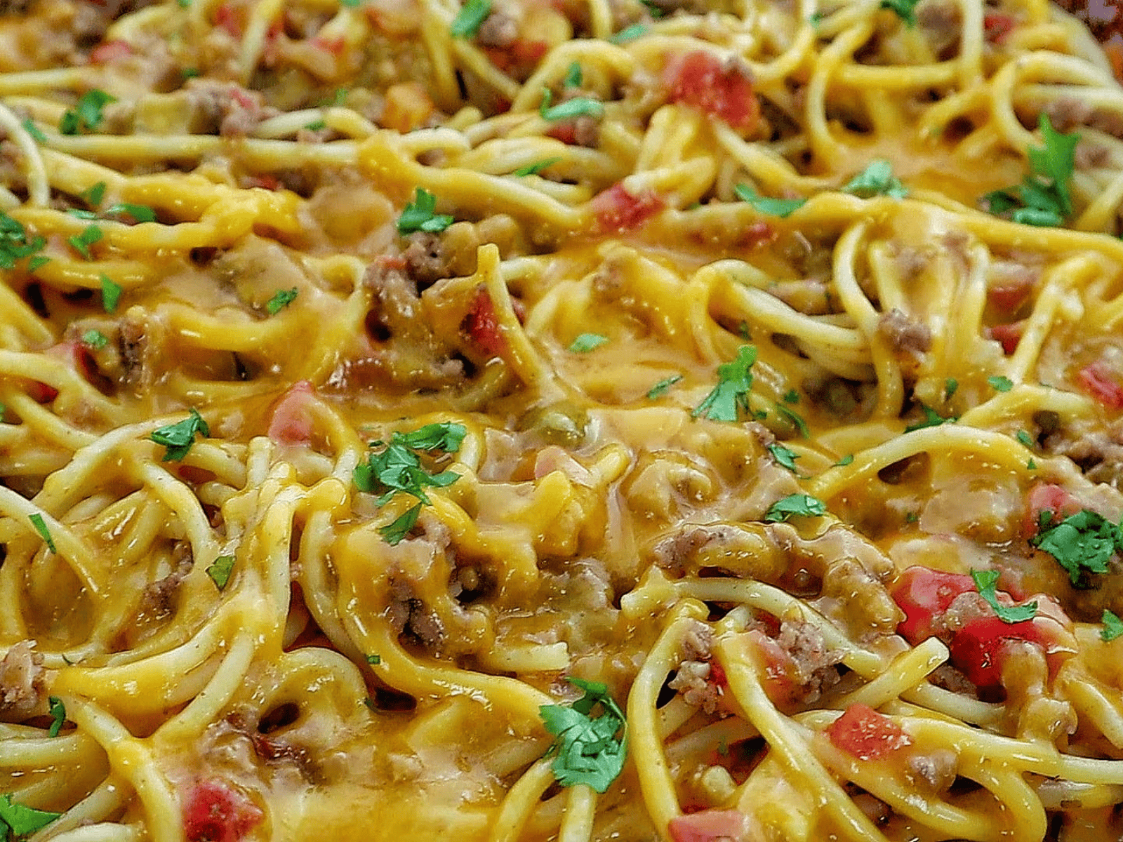 A skillet filled with cheesy taco spaghetti, featuring seasoned ground beef, spaghetti noodles, melted cheddar cheese, diced tomatoes, and garnished with chopped fresh parsley.