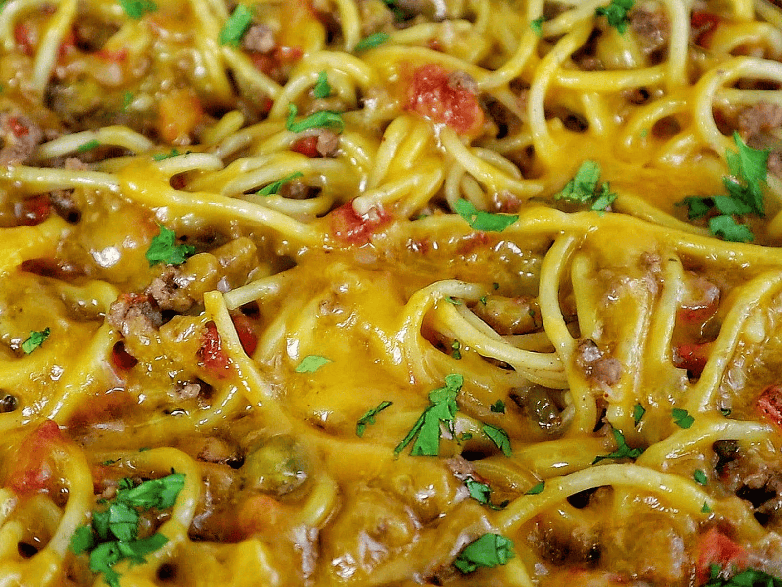 A skillet filled with cheesy taco spaghetti, featuring seasoned ground beef, spaghetti noodles, melted cheddar cheese, diced tomatoes, and garnished with chopped fresh parsley.