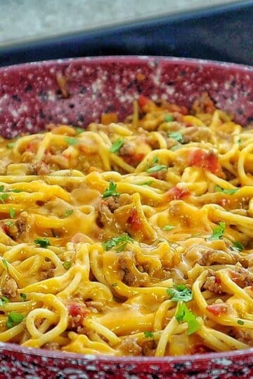 A skillet filled with cheesy taco spaghetti, featuring seasoned ground beef, spaghetti noodles, melted cheddar cheese, diced tomatoes, and garnished with chopped fresh parsley.
