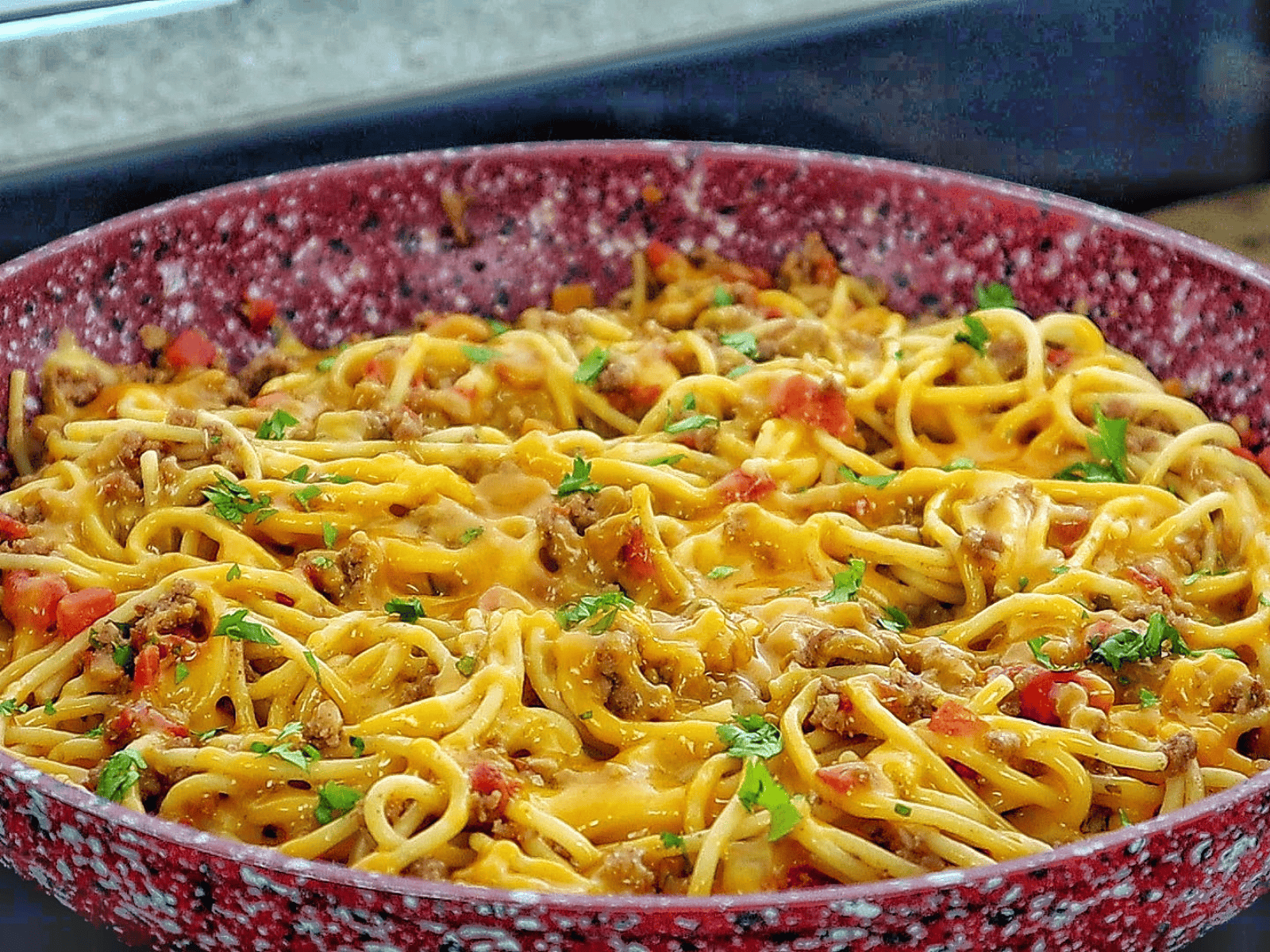 A skillet filled with cheesy taco spaghetti, featuring seasoned ground beef, spaghetti noodles, melted cheddar cheese, diced tomatoes, and garnished with chopped fresh parsley.