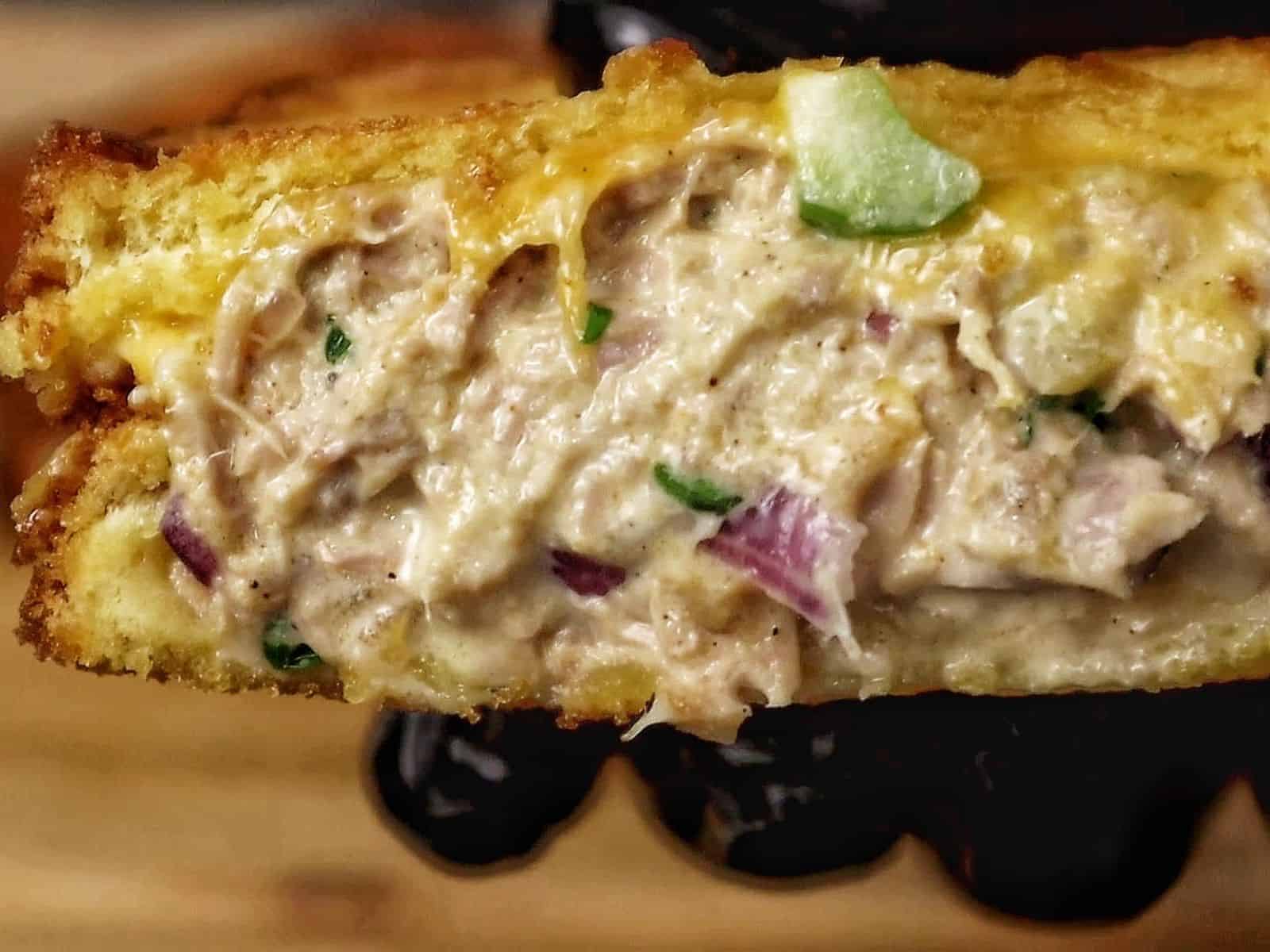 A golden-brown grilled tuna melt sandwich stacked in halves, oozing with melted cheddar cheese and filled with a creamy tuna salad mixture of tuna, onions, and celery.