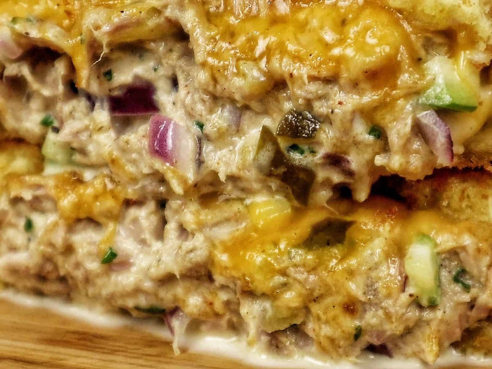 A golden-brown grilled tuna melt sandwich stacked in halves, oozing with melted cheddar cheese and filled with a creamy tuna salad mixture of tuna, onions, and celery.