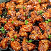 Crispy Chinese orange chicken coated in a sticky orange glaze, garnished with sesame seeds and chopped green onions in a skillet.