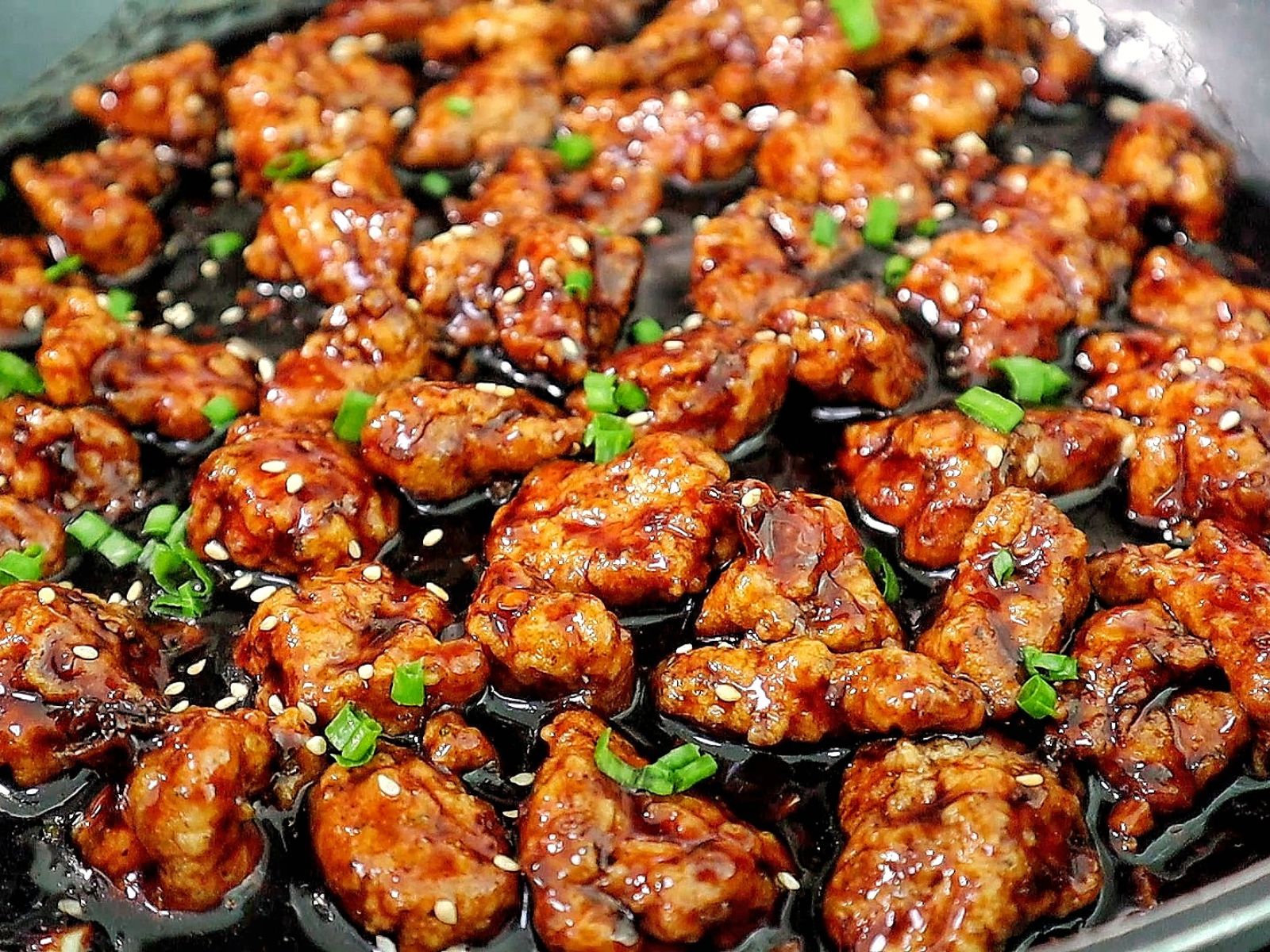 Crispy Chinese orange chicken coated in a sticky orange glaze, garnished with sesame seeds and chopped green onions in a skillet.