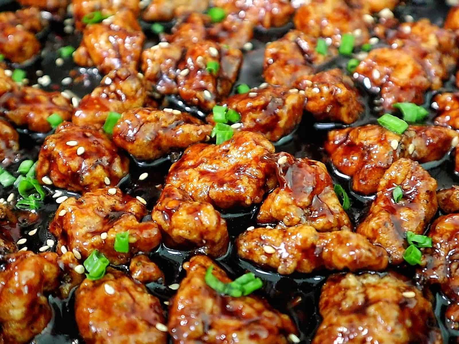 Crispy Chinese orange chicken coated in a sticky orange glaze, garnished with sesame seeds and chopped green onions in a skillet.
