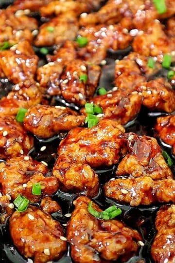 Crispy Chinese orange chicken coated in a sticky orange glaze, garnished with sesame seeds and chopped green onions in a skillet.
