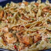A skillet of creamy white cheddar chicken pasta made with bowtie noodles, tender seared chicken chunks, and a rich cheesy sauce, garnished with parsley and a sprinkle of red pepper flakes.