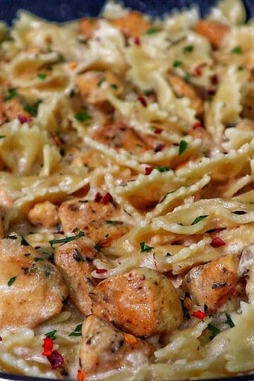 A skillet of creamy white cheddar chicken pasta made with bowtie noodles, tender seared chicken chunks, and a rich cheesy sauce, garnished with parsley and a sprinkle of red pepper flakes.