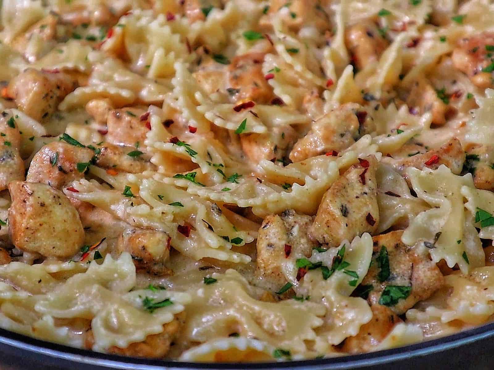 A skillet of creamy white cheddar chicken pasta made with bowtie noodles, tender seared chicken chunks, and a rich cheesy sauce, garnished with parsley and a sprinkle of red pepper flakes.