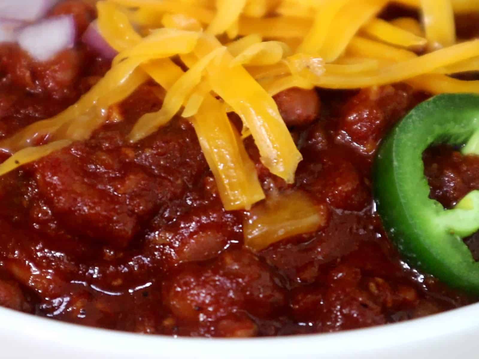 A hearty bowl of steak chili topped with shredded cheddar cheese, chopped red onions, jalapeño slices, and a dollop of sour cream, served hot and ready to eat.
