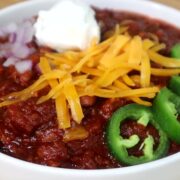 A hearty bowl of steak chili topped with shredded cheddar cheese, chopped red onions, jalapeño slices, and a dollop of sour cream, served hot and ready to eat.
