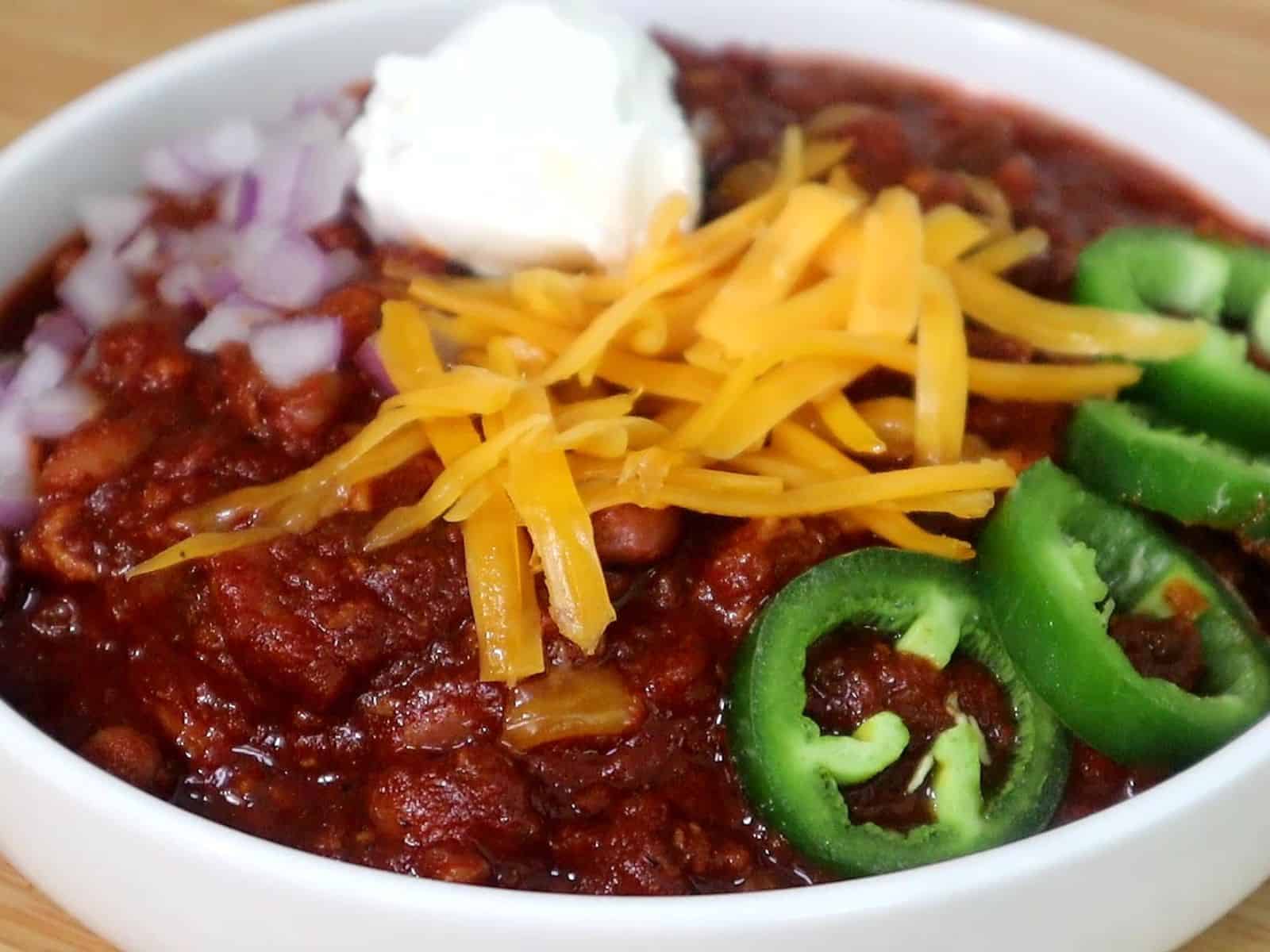A hearty bowl of steak chili topped with shredded cheddar cheese, chopped red onions, jalapeño slices, and a dollop of sour cream, served hot and ready to eat.