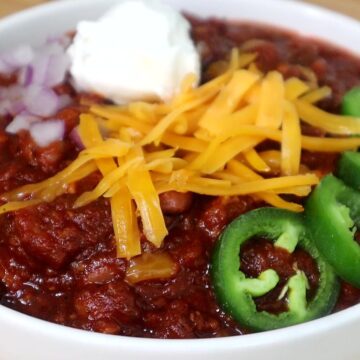 A hearty bowl of steak chili topped with shredded cheddar cheese, chopped red onions, jalapeño slices, and a dollop of sour cream, served hot and ready to eat.