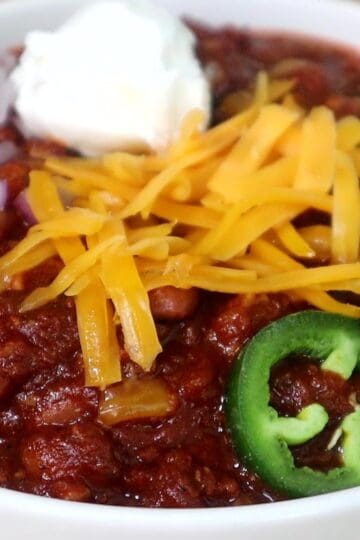A hearty bowl of steak chili topped with shredded cheddar cheese, chopped red onions, jalapeño slices, and a dollop of sour cream, served hot and ready to eat.
