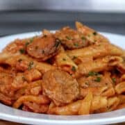 A skillet filled with penne pasta coated in creamy tomato sauce mixed with sliced smoked sausage, colorful bell peppers, and fresh herbs.