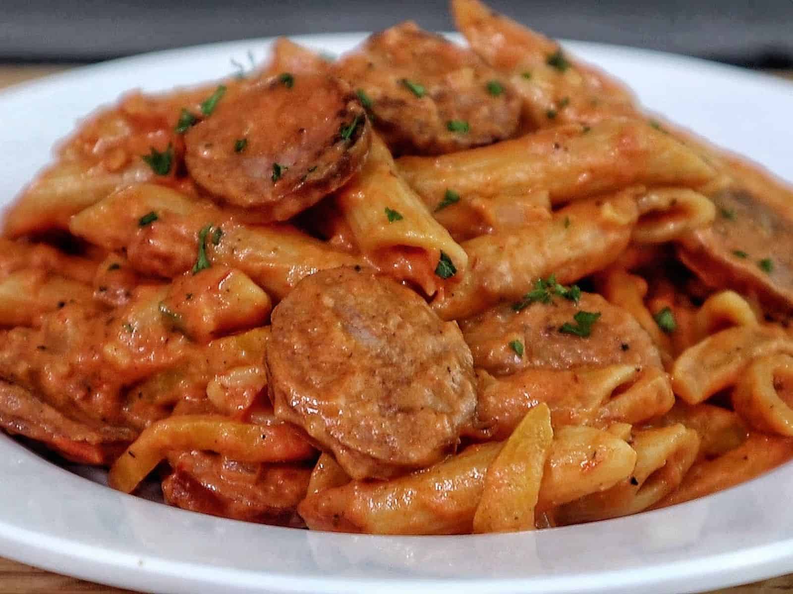 A skillet filled with penne pasta coated in creamy tomato sauce mixed with sliced smoked sausage, colorful bell peppers, and fresh herbs.