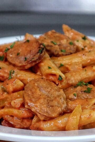 A skillet filled with penne pasta coated in creamy tomato sauce mixed with sliced smoked sausage, colorful bell peppers, and fresh herbs.