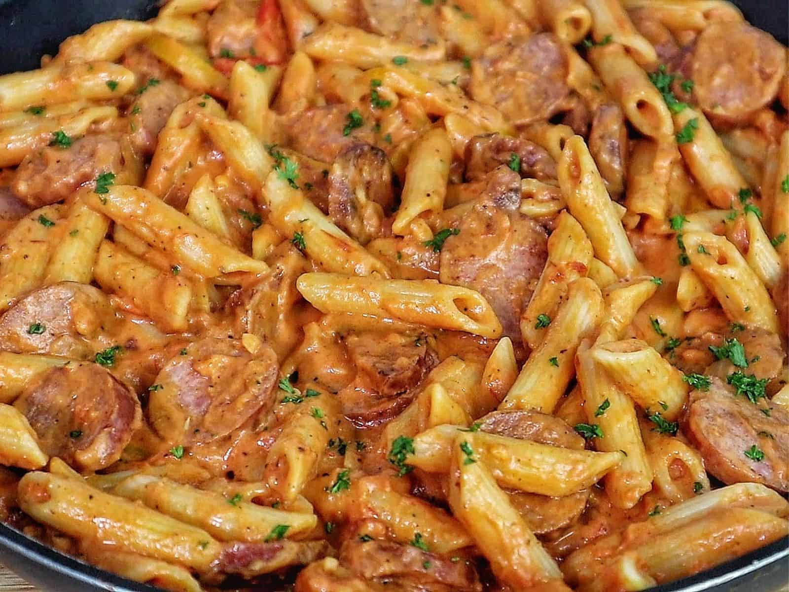 A skillet filled with penne pasta coated in creamy tomato sauce mixed with sliced smoked sausage, colorful bell peppers, and fresh herbs.