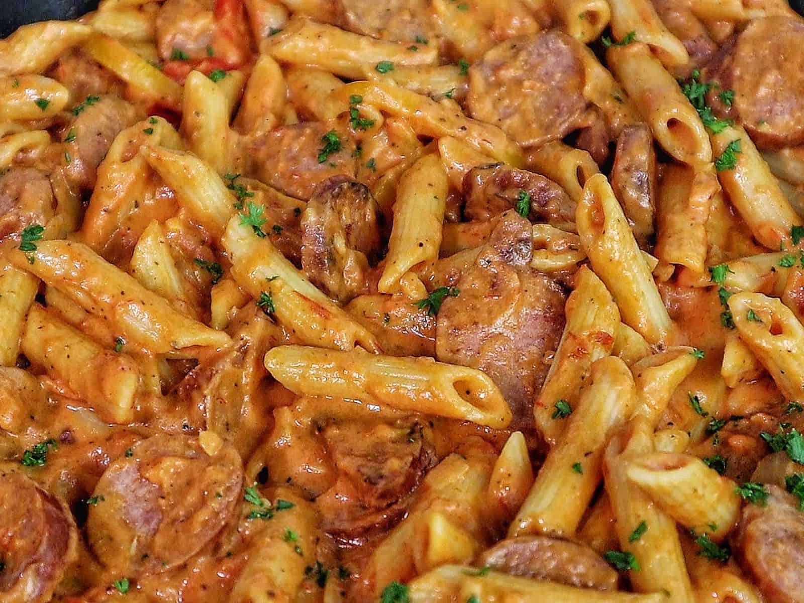 A skillet filled with penne pasta coated in creamy tomato sauce mixed with sliced smoked sausage, colorful bell peppers, and fresh herbs.